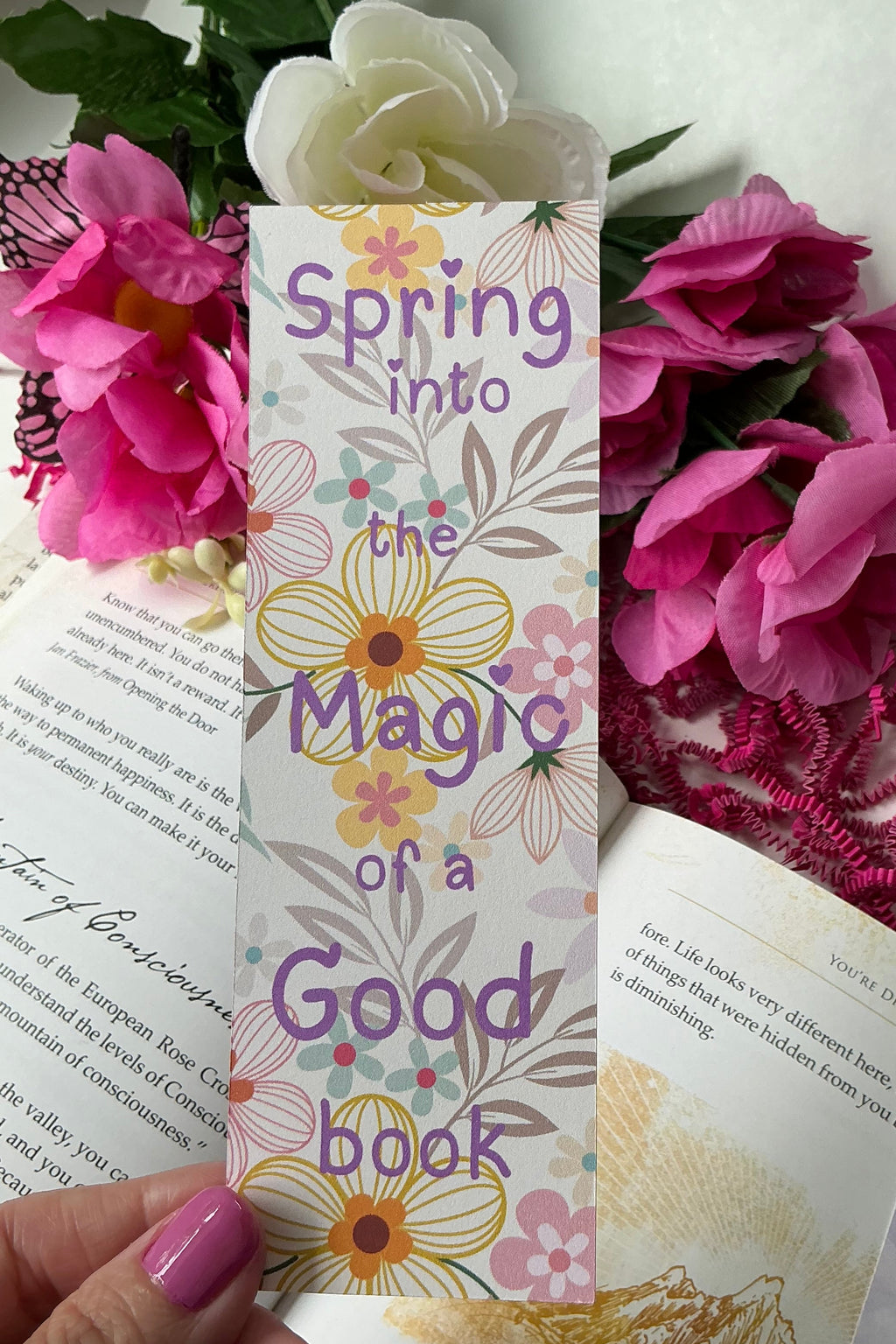 Colorful bookmarks with floral designs held over a book and flowers on a white surface