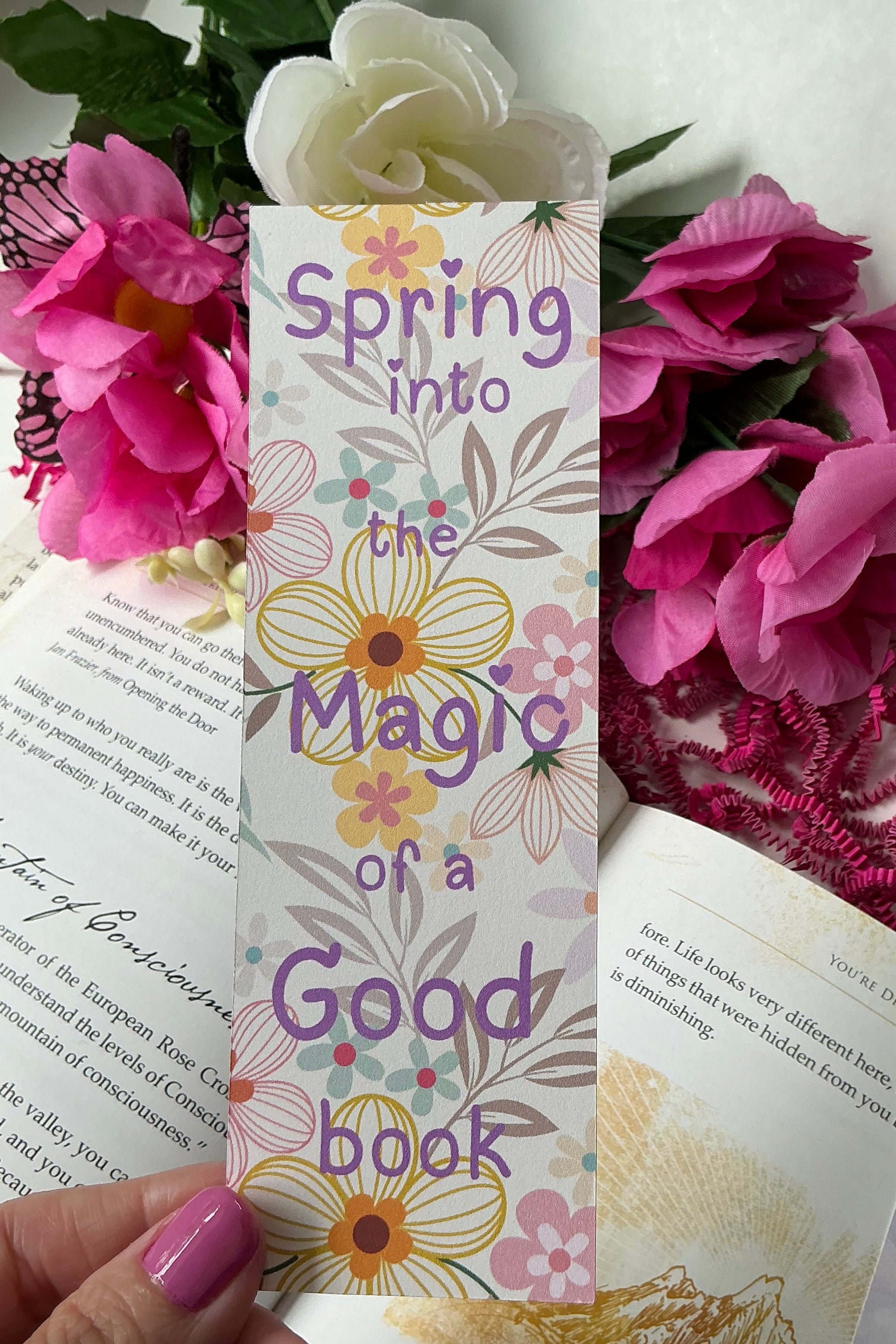 Colorful bookmarks with floral designs held over a book and flowers on a white surface
