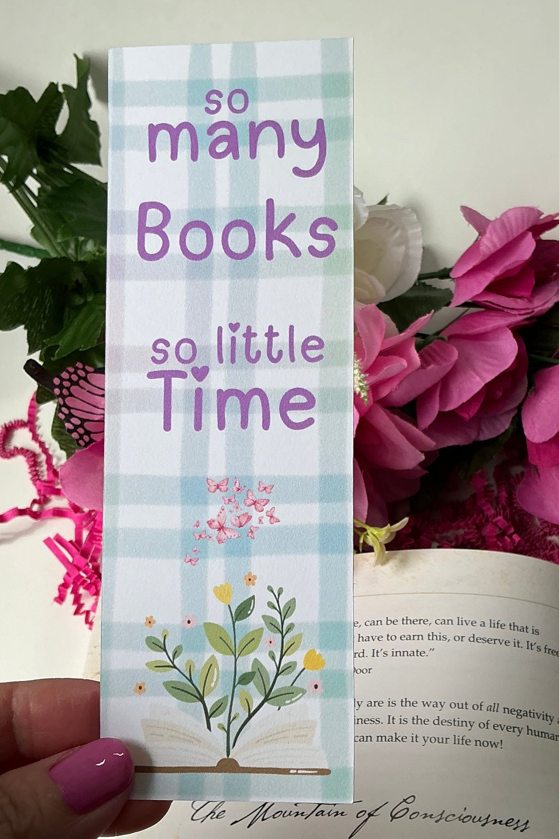 Hand holding a bookmark with floral design and text over an open book with flowers.